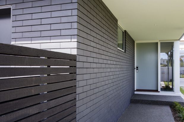 A modern residential entrance viewed from an exterior walkway. The facade is clad in dark grey horizontal brickwork with a narrow horizontal window, while a dark timber slat fence leads toward a glazed white-framed entry door. The covered porch and textured dark paving create a restrained, contemporary composition.