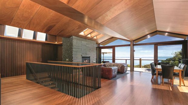 A warm, open-plan contemporary interior with a dramatic timber-lined pitched ceiling and extensive glazing overlooking a body of water. The space includes a living area with lounge chairs, a dining area, a dark timber kitchen or stair-side joinery zone, a stone-clad fireplace feature wall, and a black vertical-slat balustrade around a stair opening. Skylights and clerestory windows bring in abundant natural light.