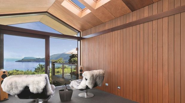 A compact, richly timber-lined interior overlooking a bay and green headland. Floor-to-ceiling glazing opens to a balcony, while skylights bring daylight through the angled timber ceiling. Two sculptural white sheepskin lounge chairs, compact speakers, and a small side table create a warm, distinctive retreat-like seating space.
