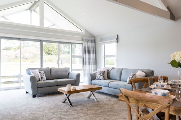 Bright, spacious living room with a vaulted ceiling, exposed timber beam, high clerestory glazing, and expansive sliding glass doors. The room features two grey sofas, a timber coffee table, a timber dining table with cross-back chairs, neutral carpet, and patterned curtains. The overall design is relaxed, contemporary, and airy.
