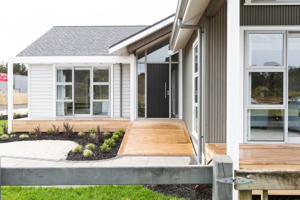 Exterior view of a contemporary single-storey building with interconnected gabled volumes, grey horizontal cladding, dark vertical siding, white-framed windows, and a timber deck and ramp leading to the entry. The landscaped foreground includes a gravel garden, low planting, concrete edging, and a timber boundary structure.