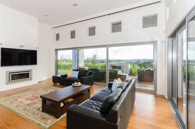 Bright contemporary living room with a double-height ceiling, polished timber flooring, black leather sofas, a dark timber coffee table, wall-mounted television and built-in fireplace. Floor-to-ceiling sliding glass doors open to a landscaped outdoor terrace, while high-level windows bring in additional light and views.