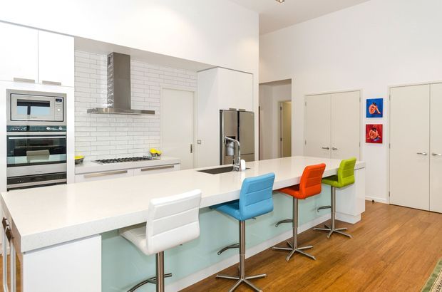 Bright contemporary kitchen with a large white island and integrated breakfast bar, four colourful bar stools, white cabinetry, stainless-steel appliances, a white subway-tile splashback, and warm timber flooring. The space has a clean, open layout with concealed storage and a small hallway visible beyond.