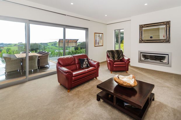Bright contemporary living room with white walls, recessed ceiling lights, beige carpet, and large sliding glass doors opening to a furnished balcony with a green rural landscape beyond. The room features deep red leather sofas and armchair, a dark timber coffee table, a wall-mounted gas fireplace, and an ornate framed mirror.