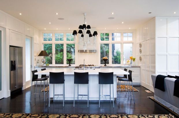 Bright, spacious white kitchen with extensive built-in cabinetry, a large central island with a white benchtop, stainless-steel appliances, dark timber flooring, and multiple black bar stools. The room features large multi-pane windows, a decorative black chandelier, recessed lighting, and a glimpse of upholstered seating at the right edge.
