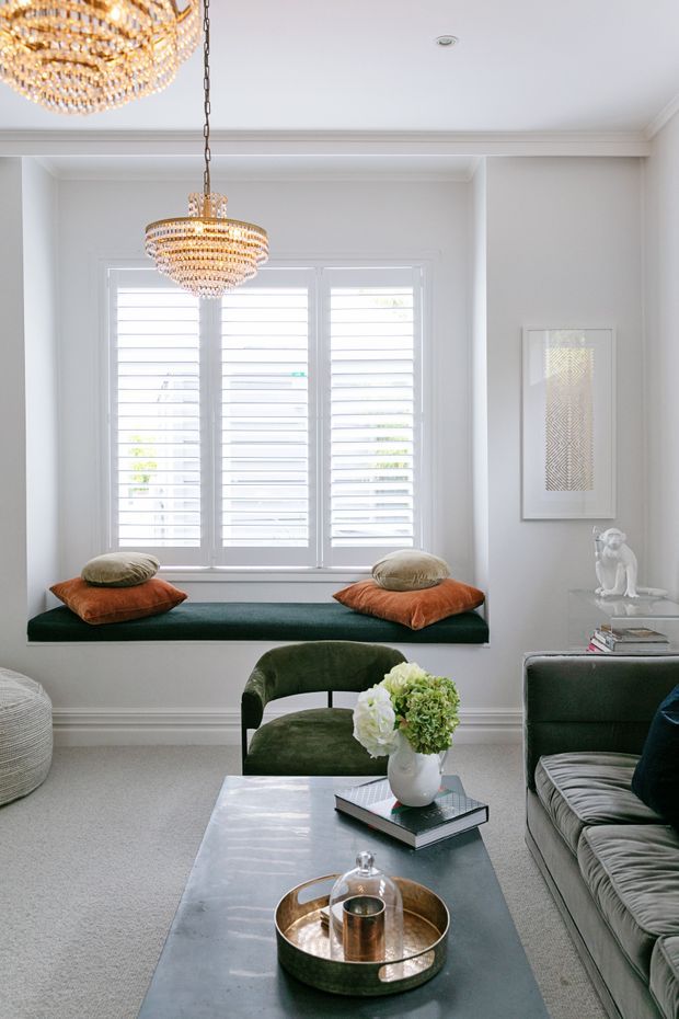 Bright, refined living room with white walls and trim, a large shuttered window with a built-in deep green window seat, velvet green seating, rust-orange cushions, and an elegant marble-topped coffee table. Crystal and gold pendant chandeliers add a luxe accent, while framed artwork and carefully arranged accessories complete the room.