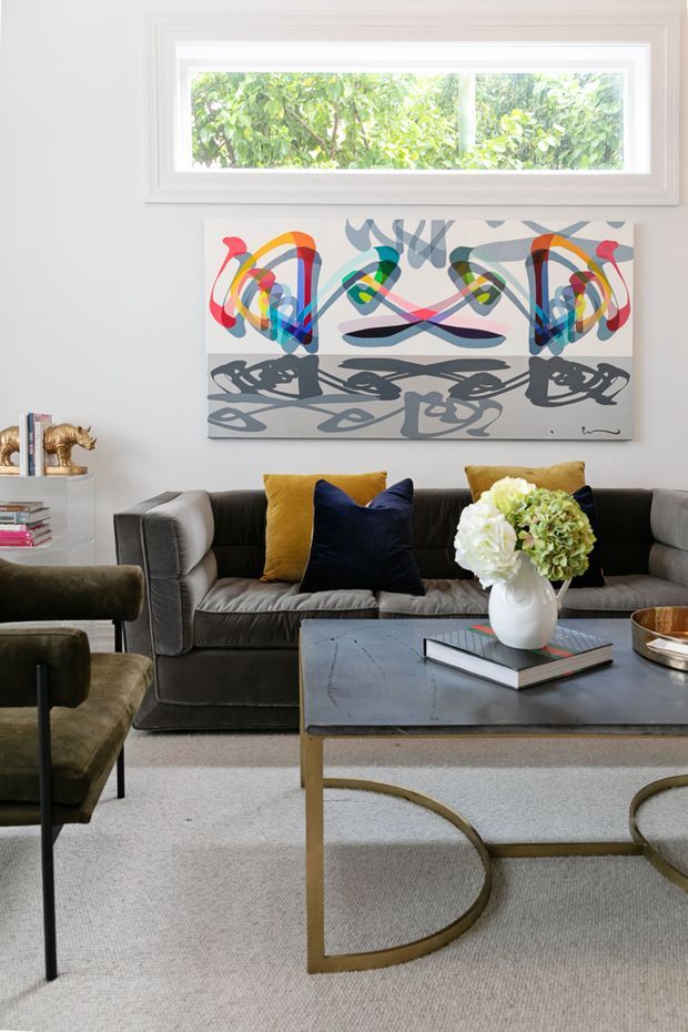 A refined living room with a charcoal grey sofa, mustard and navy cushions, a dark rectangular coffee table with curved brass-toned legs, and sculptural olive armchairs. A large colourful abstract artwork hangs beneath a wide horizontal window, while a white vase of flowers, books, and metallic accessories add decorative detail. The room has pale walls and a light neutral carpet, creating a polished, art-led contemporary interior.