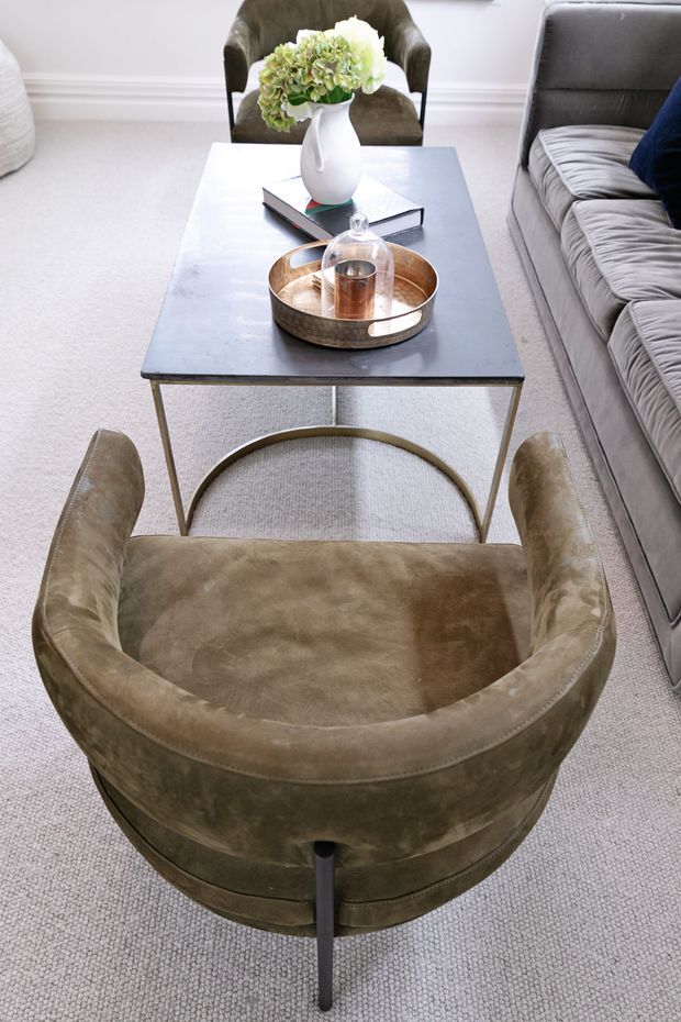 A bright, refined living room vignette featuring a deep olive-brown velvet barrel armchair in the foreground, a grey upholstered sofa to the right, and a rectangular dark coffee table with a slim metal frame and curved stretcher. The table is styled with a metallic tray, candle, books, and a white vase of hydrangeas. Pale carpet, white walls, and restrained neutral furnishings create an elegant, softly layered interior.