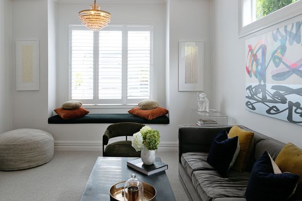 Bright, refined living room with a built-in window seat beneath plantation shutters, a grey sofa with jewel-toned cushions, a low marble or stone coffee table, sculptural pendant lighting, and colourful abstract wall art. The room has pale walls, carpeted flooring, and a calm contemporary composition with traditional window detailing.