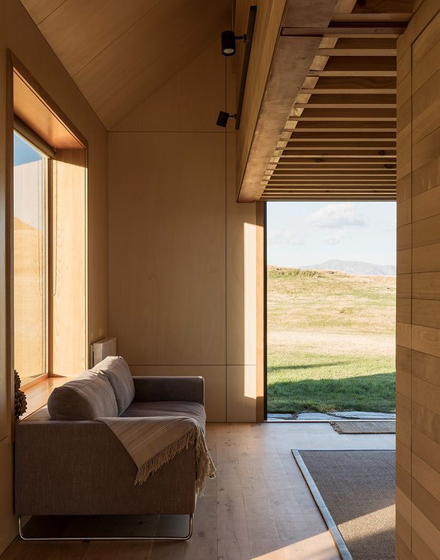 A compact, warm-toned timber-lined sitting room or cabin interior with a low upholstered sofa beside a large window. The room has timber wall and ceiling panels, a timber floor, minimal black track lighting, and an open glazed doorway leading to a grassy rural landscape with distant hills. Exposed timber slats form a projecting overhead structure near the doorway, adding architectural texture. The atmosphere is calm, natural, and softly lit.
