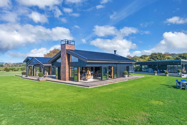 A contemporary single-storey house set within a broad manicured lawn. The building has dark metal gabled roofs, extensive floor-to-ceiling glazing, dark vertical cladding, and a prominent brick chimney or fireplace tower. A covered glazed connection extends to the right, with outdoor furniture and low planting around the paved terrace. The image is photographed from the landscaped grounds under a bright, partly cloudy sky.