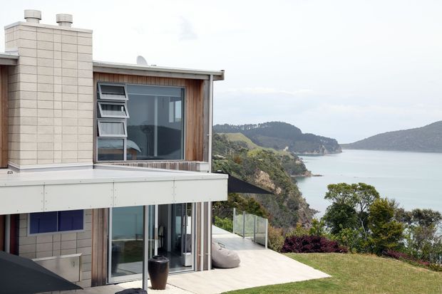 Modern multi-level coastal home on a sloping site, shown from the exterior with pale tiled wall cladding, warm vertical timber detailing, extensive glazing, and a broad light-coloured terrace overlooking a sheltered bay and wooded hills. The building has a flat roof, projecting upper floor, glass balustrades, and clean rectilinear forms.
