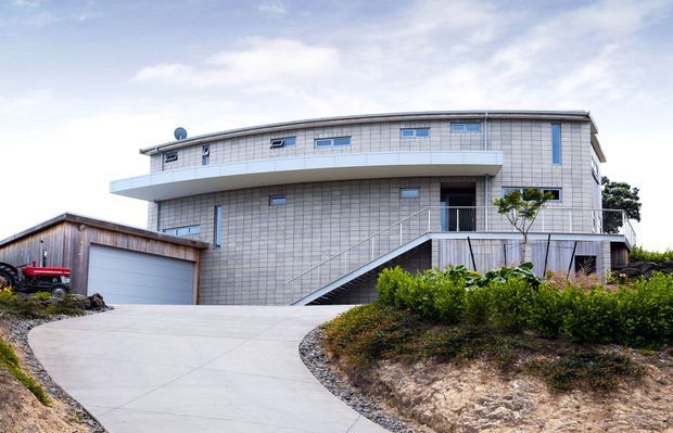Exterior view of a large contemporary multi-level residence on a sloping site. The building has a curved, rounded facade clad in small pale tiles or panels, narrow horizontal windows with blue accents, a broad projecting balcony, and an external stair rising to an upper entry. A separate garage with a wide sectional door sits to the left, while landscaped garden beds and a curved concrete driveway occupy the foreground.