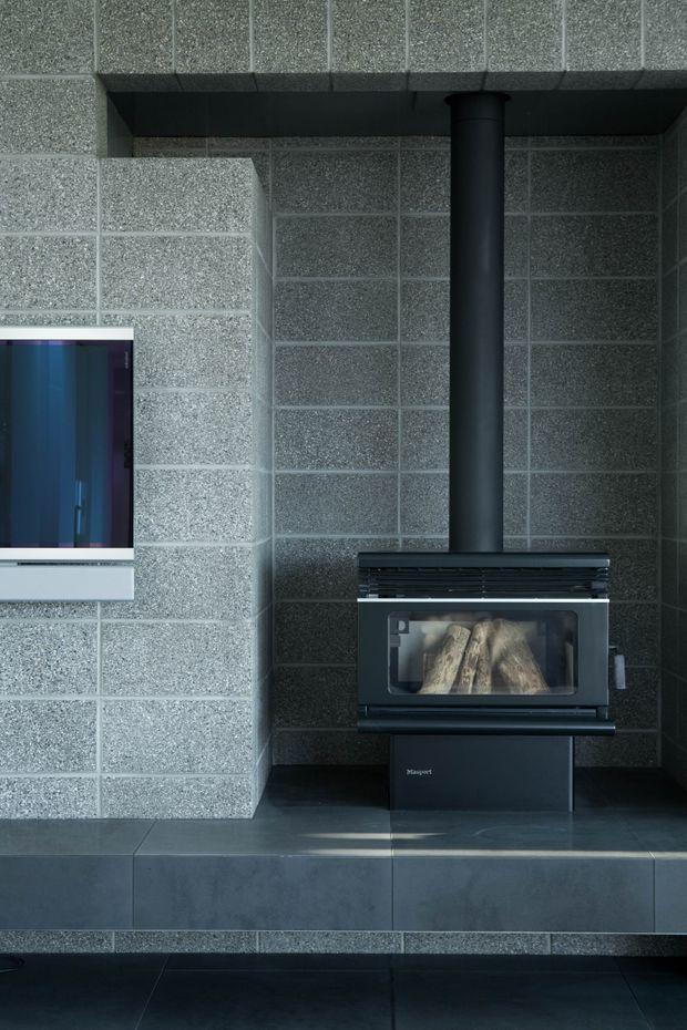 Close-up interior view of a contemporary fireplace installation. A black freestanding wood-burning stove with visible logs and a tall black flue sits on a dark tiled hearth, surrounded by grey speckled masonry block walls. Part of a wall-mounted television is visible at left. The space has a dark, cool-toned, utilitarian finish.