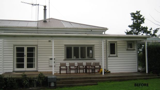 Photograph of the front or rear exterior of a low-rise traditional house, showing pale horizontal weatherboard cladding, a corrugated hipped roof with chimney and antenna, white-trimmed windows and doors, and a covered porch/deck with outdoor seating. The image is labelled “BEFORE,” indicating a pre-renovation condition.