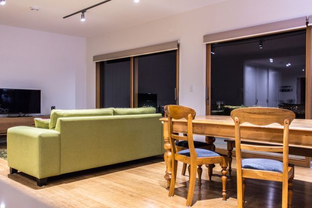 Open-plan living and dining area with a pale green sofa, timber dining table and chairs, light timber flooring, recessed and track lighting, and large dark-framed windows or glazed doors with roller blinds. The room has a simple contemporary fit-out and warm timber furnishings.