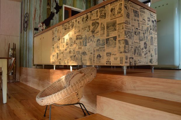 A warm, eclectic mid-century-inspired interior featuring a raised timber platform and extensive timber wall and floor finishes. A low cabinet with retro newspaper-print cladding dominates the scene, accompanied by a woven wicker lounge chair with a black metal frame. The room also includes distressed patterned wall panels, a frosted glass door or partition, and vintage decorative elements.