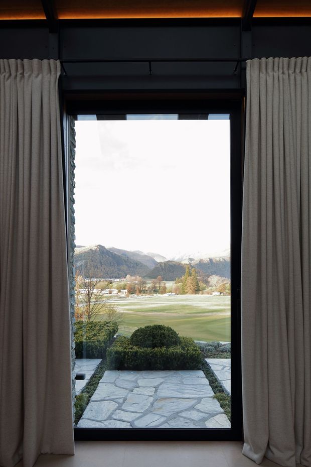 A tall, narrow floor-to-ceiling window framed in dark metal is flanked by full-length pleated curtains. Through the glazing, a pale stone pathway leads across clipped garden hedges toward an open grassy landscape and dramatic mountain ranges. The interior is dim and refined, with warm linear ceiling lighting and dark structural framing contrasting with the bright alpine view.