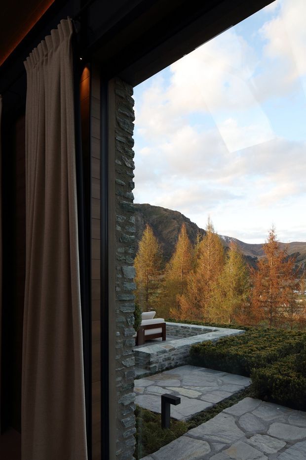 A dramatic view from inside a dark, refined interior through a tall glazed opening toward a landscaped stone terrace and mountainous autumn landscape. Heavy floor-to-ceiling taupe curtains frame the opening, while a rugged stacked-stone wall, dark window framing, pale irregular stone paving, clipped shrubs, and a simple outdoor lounge chair create a connection between the interior and the alpine setting.