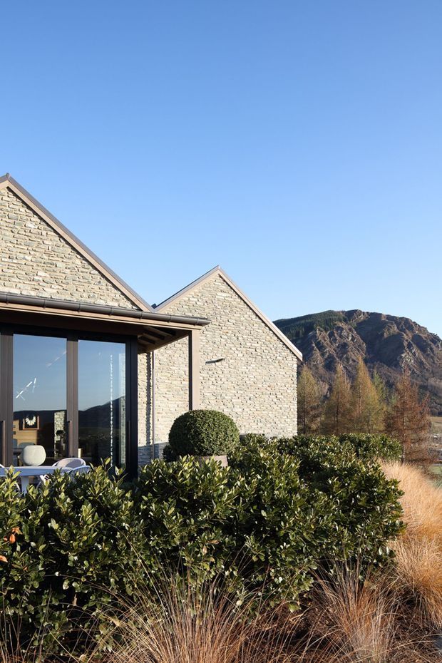 Exterior view of a contemporary gabled building clad in pale stacked stone, with large dark-framed glazed doors and windows opening toward landscaped planting. Ornamental shrubs and grasses surround the structure, with rugged mountainous terrain visible in the background under a clear blue sky.