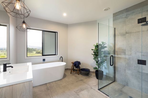 Bright, spacious contemporary bathroom featuring a freestanding white bathtub, double vanity with pale wood cabinetry, a large frameless glass shower enclosure with grey concrete-look wall tiles, and black tapware and geometric pendant lights. A rural landscape is visible through the windows.