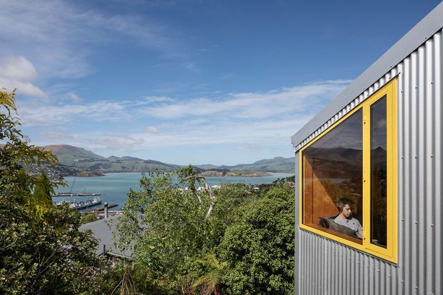 Exterior view of a contemporary corrugated-metal-clad building with a sharply sloping roof edge and a prominent yellow-framed window. The building overlooks a blue harbour or inlet surrounded by hilly terrain, with greenery in the foreground. A person is visible through the window, but the exterior envelope is the main subject.