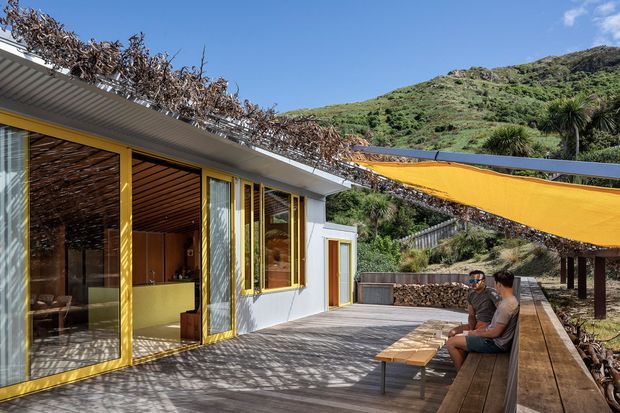 Bright exterior view of a modern single-storey dwelling opening onto a timber deck. The building has white walls, extensive yellow-framed glazing and doors, a low corrugated roof, and warm timber-lined interior surfaces visible through the open sliding doors. A large orange shade sail covers part of the outdoor seating area, with a green hillside landscape beyond.