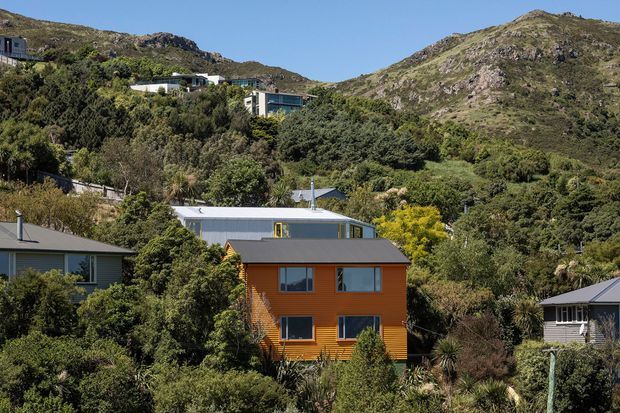 Exterior view of a compact orange timber-clad house nestled in dense vegetation on a steep, mountainous suburban site. The house has a simple two-storey rectangular form, dark pitched roof, and large rectangular windows. Other homes are visible among the trees and rugged hills rise directly behind the neighbourhood under a clear blue sky.