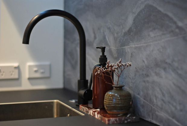 Close-up of a contemporary kitchen or utility sink area featuring a tall matte black gooseneck tap, dark undermount sink, grey marble-look splashback, and a dark stone or composite benchtop. An amber soap dispenser and decorative vase sit on a small tray beside the tap.