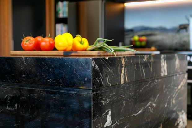 Close-up view of a contemporary kitchen island with a dramatic dark veined stone finish and a warm timber benchtop. Fresh vegetables are arranged on the counter, while dark cabinetry, integrated lighting, and a reflective window or splashback are visible in the background.