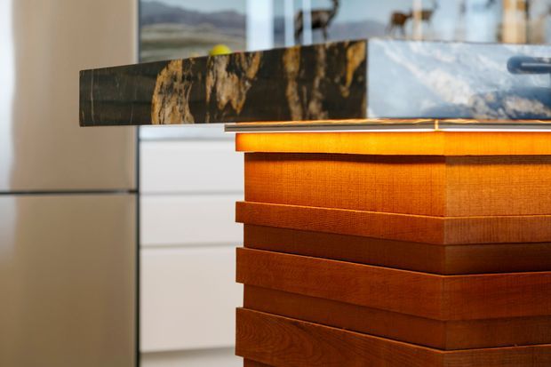 Close-up architectural detail of a kitchen island featuring richly grained warm timber cabinetry with layered horizontal drawer fronts and a dark, dramatic natural-stone benchtop. White cabinetry and reflective glazing appear in the background.