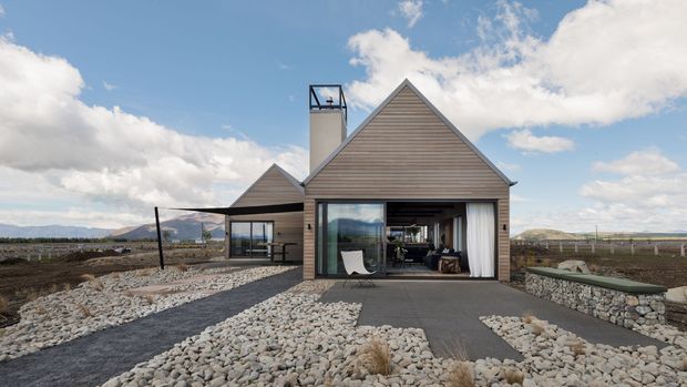 Single-storey contemporary rural home with a prominent steep gabled form, horizontal timber-look cladding, broad glazed garage or living opening, and a smaller side wing. The house sits in a sparsely planted, rocky landscape with gravel paths and low stone retaining walls, beneath a bright partly cloudy sky. A dark metal-framed canopy connects to the side of the building.