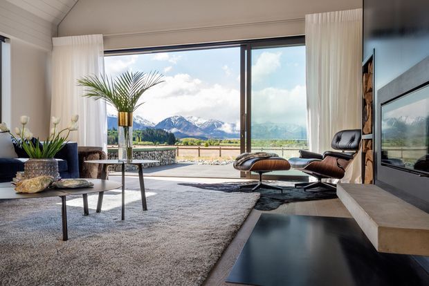 A bright, refined living room with expansive floor-to-ceiling glazing overlooking a mountain landscape. The room features a contemporary fireplace and media wall, a classic leather lounge chair with ottoman, dark blue seating, a large pale textured rug, glass and metal coffee tables, indoor palms, and sheer white curtains. The ceiling and walls include subtle horizontal timber-style panelling.
