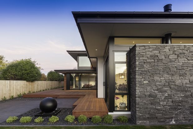 Exterior view of a contemporary multi-level residence with broad low-pitched roof overhangs, dark-framed glazing, timber decking, and a prominent stacked-stone privacy wall. A landscaped entry garden and paved driveway occupy the foreground, with warm interior furnishings visible through the windows.