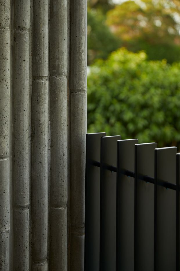 Close-up architectural detail showing a tall ribbed, board-formed concrete wall or column beside a dark matte metal privacy screen or balustrade. Dense blurred greenery is visible beyond the opening, creating a strong contrast between heavy textured construction and soft natural landscape. The image is vertically composed with a muted, moody palette.