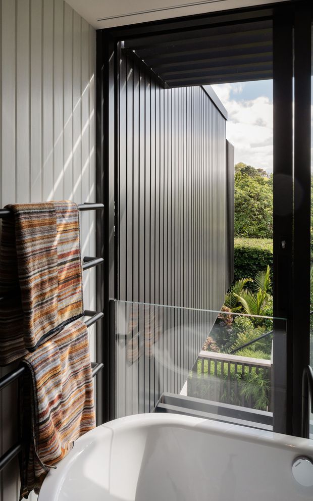 A contemporary bathroom featuring a white freestanding bathtub positioned beside full-height glazing. Black-framed openings, dark vertically ribbed exterior cladding, a glass balustrade, and a lush garden view create a strong indoor–outdoor connection. A striped towel hangs on a dark heated towel rail in the foreground.