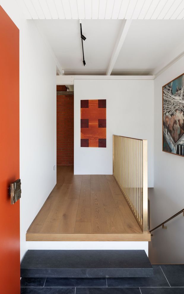 A bright contemporary residential stair landing and entry transition. The space features a warm timber landing floor, dark tiled lower steps, crisp white walls and ceiling with exposed linear detailing, a vivid orange-red door, a slim black track light, and a gold-toned vertical balustrade. Abstract artwork and a red brick wall add visual texture.