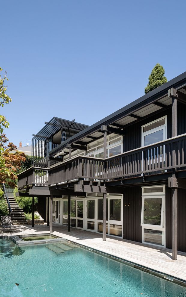 Exterior view of a contemporary multi-level dark-clad residence with extensive timber decking, elevated balconies, vertical slatted screening and broad glazed doors and windows. A swimming pool occupies the foreground, with stairs and landscaped planting along the side of the house under a clear blue sky.