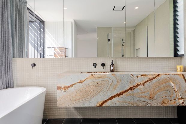 Contemporary bathroom featuring a freestanding white bathtub, wall-mounted black tapware, a large mirrored wall, glass shower enclosure, and a striking veined stone vanity with integrated basin. Small textured wall tiles, dark floor tiles, and soft grey curtains create a refined, high-contrast finish.