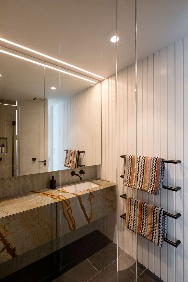 A refined contemporary bathroom featuring a floating stone vanity with an integrated basin, dark tiled lower cabinetry, a large mirrored wall, and a wall-mounted multi-rail towel warmer. Vertical white wall panelling and discreet linear ceiling lighting create a clean, elongated composition. A textured small-format tiled splashback and dramatic veined natural stone add visual contrast.