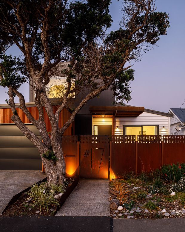 Twilight street-facing view of a contemporary residential facade and entry. A mature sculptural tree dominates the foreground, with a concrete path leading through a rust-coloured perforated metal gate and fence. The house features horizontal light cladding, dark grey metal elements, warm concealed and wall lighting, timber-toned screening, and a mix of flat and sloping roof forms. Low-maintenance native-style planting and gravel landscaping frame the entrance.