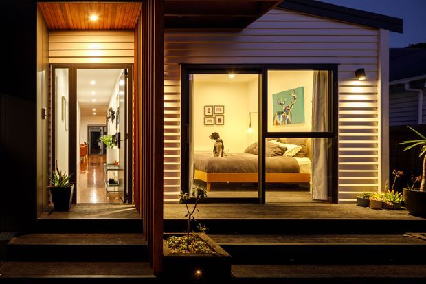 Night-time exterior view of a compact contemporary dwelling, featuring a warmly lit recessed entry and a bedroom visible through large black-framed glazing. Horizontal light-coloured cladding, dark timber decking and steps, timber-lined soffits, and low garden planters define the facade. The bedroom contains a bed and framed artwork, while a long illuminated hallway is visible through the entry.