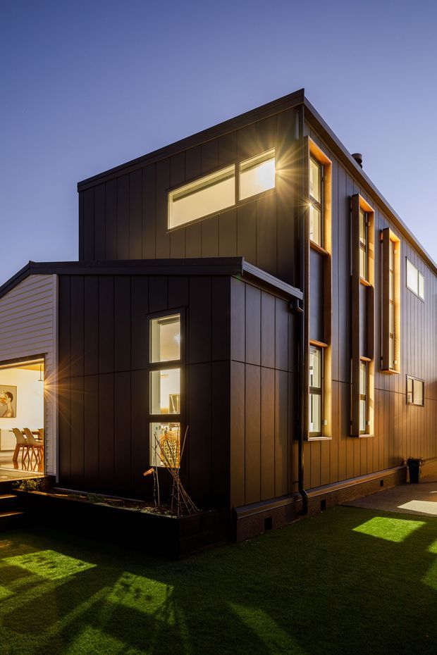 Twilight exterior view of a modern multi-storey residential building with dark vertical panel cladding, warm timber-framed windows, projecting window surrounds, and a low connecting wing. The building forms a compact angular mass around a small lawn and planted edge, with warm interior lighting visible through the windows.