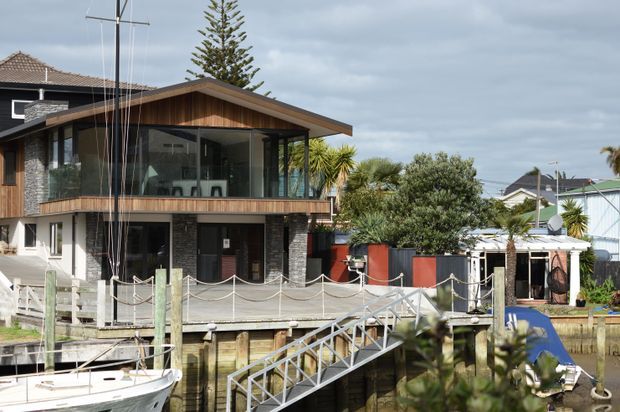Exterior view of a contemporary multi-level waterfront home with a prominent timber-clad upper pavilion, extensive dark-framed glazing, exposed balcony, and a gabled roof. A marina-style timber deck, boat ramp, and dock occupy the foreground, with neighboring homes and coastal vegetation surrounding the property.