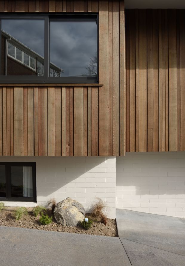 Close-up exterior view of a contemporary building facade combining vertical natural timber cladding with white-painted masonry at ground level. Dark-framed windows reflect the surrounding structure and sky. A simple concrete path, gravel planting bed, grasses, and a large boulder sit along the base of the building.