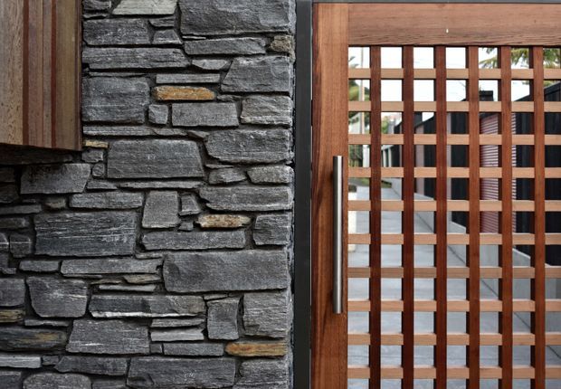 Close-up architectural detail of a contemporary entrance, showing rough stacked dark stone wall cladding beside a warm timber-framed glazed door with a square timber lattice screen and vertical metal pull handle. A courtyard or exterior area is visible beyond the door.