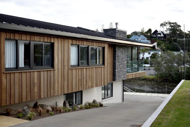 Exterior architectural view of a contemporary multi-level house on a sloped site. The building features vertical timber cladding, a dark pitched metal roof, large dark-framed windows, a stone-clad chimney or feature wall, and a pale rendered lower level. A driveway and landscaped foreground are visible, with neighbouring hillside houses in the background.
