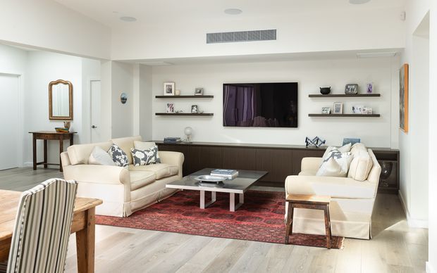 Bright, spacious contemporary living room with two cream sofas, a dark built-in media wall and floating display shelves, a wall-mounted television, coffee table, patterned red rug, and pale timber flooring. A small entry or hallway area is visible to the left.