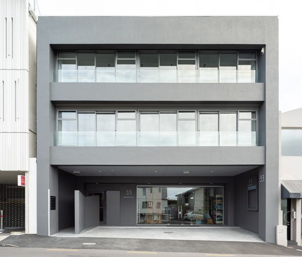 Street-level exterior photograph of a contemporary multi-storey residential or mixed-use building with a dark grey rendered facade, three stacked levels, broad recessed balconies, extensive glazed openings, and a covered ground-floor entry or garage-like frontage. The design has clean rectilinear lines and aluminium-framed glazing.