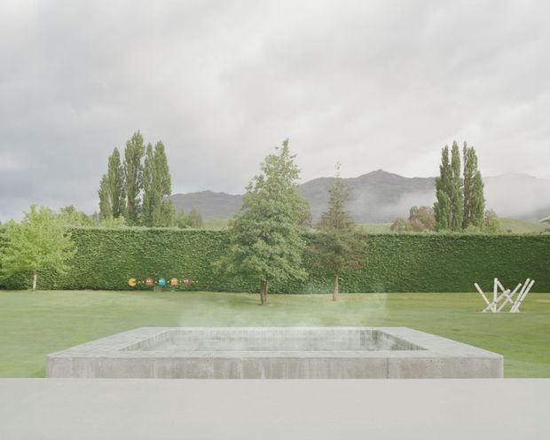 A minimalist landscaped outdoor area viewed from a broad pale concrete terrace, featuring a rectangular in-ground concrete pool or water basin in the foreground. A manicured lawn, tall clipped hedge, mature trees, and distant misty mountains sit beneath an overcast sky. The restrained composition and raw concrete elements create a serene, contemporary atmosphere.